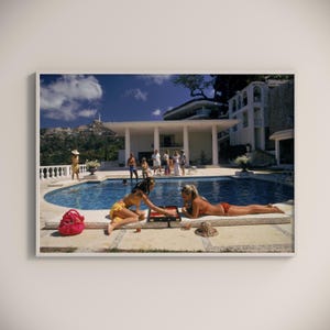 May include: Two people in swimsuits play a board game by a pool at a luxury villa. The pool is surrounded by a white patio and a lush green lawn. The villa has a white exterior with large windows and a balcony. The sky is blue with white clouds.