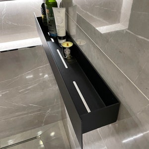 May include: A black metal shelf with two white dividers. The shelf is mounted on a gray tiled wall and contains a bottle of liquid, a tube of lotion, and a candle.