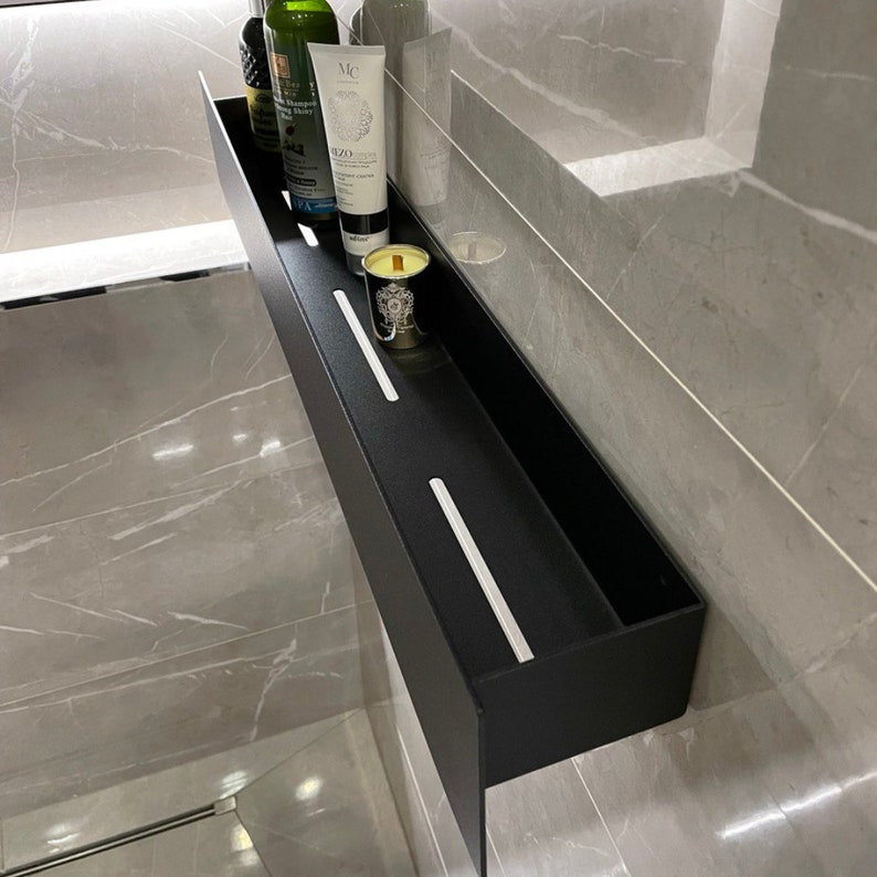 May include: A black metal shelf with two white dividers. The shelf is mounted on a gray tiled wall and contains a bottle of liquid, a tube of lotion, and a candle.