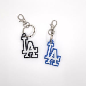 May include: Two keychains featuring the Los Angeles Dodgers logo, one in black and white, the other in blue and white. Each keychain has a silver-colored clasp for easy attachment to keys or bags. The logos are in a bold, block letter style.
