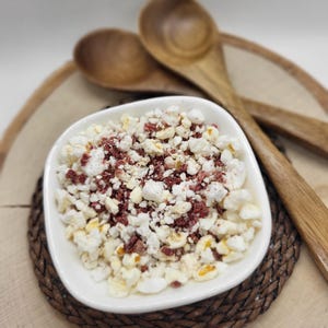 May include: A white bowl filled with popcorn and red sprinkles, sitting on a woven brown placemat. Two wooden spoons are in the background.
