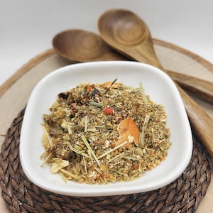 May include: A white bowl filled with a blend of dried herbs, flowers, and fruit peels. The bowl is sitting on a woven brown mat with two wooden spoons beside it.