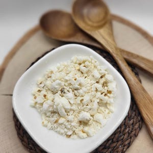 May include: A white bowl filled with crushed popcorn on a brown woven placemat. Two wooden spoons are in the background.