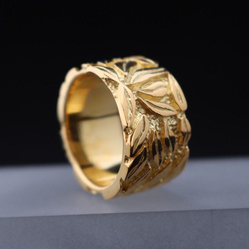 May include: A gold ring with a textured design of leaves. The ring is wide and has a smooth inner band.
