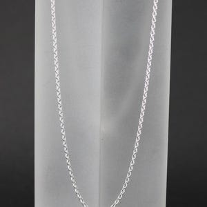 Könnte beinhalten: Eine silberfarbene Halskette mit einem filigranen, verbundenen Design. Die Halskette wird vor einem Hintergrund aus mattiertem Glas präsentiert, was einen Kontrast zum dunklen Hintergrund erzeugt. Die Kette ist etwa 50 cm lang.