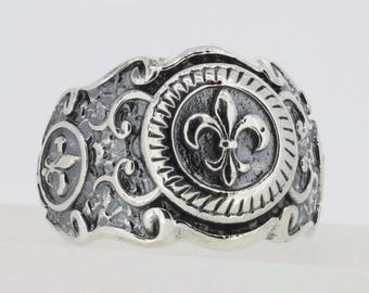 May include: A silver ring with a fleur-de-lis design. The ring has a textured surface and a detailed design.