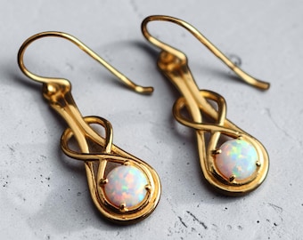 May include: A pair of gold-toned drop earrings featuring a polished metal infinity loop design. Each earring is set with a single round, iridescent white opal cabochon at the base, secured by four small prongs, and finished with a curved hook wire.