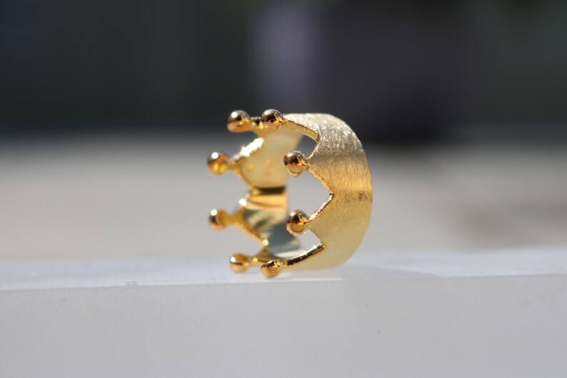 May include: A gold crown ring with a brushed finish. The ring is designed with a simple crown shape and five points.