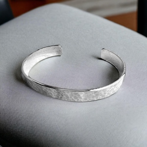 May include: A silver cuff bracelet with a hammered texture. The open-ended bracelet is made of a shiny, reflective metal. The bracelet is shown on a gray surface.