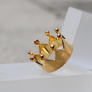 May include: A gold crown ring with a textured finish. The ring is designed with a simple, classic crown shape.