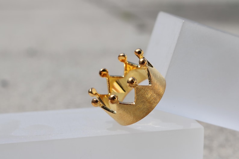 May include: A gold crown ring with a textured finish. The ring is designed with a simple, classic crown shape.