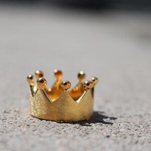 May include: A gold-colored crown ring with a textured finish. The crown has five points and is made of metal.