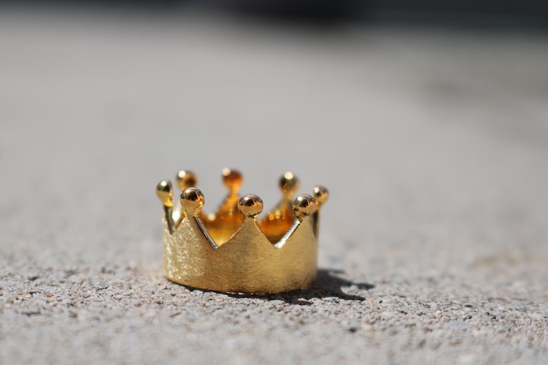 May include: A gold-colored crown ring with a textured finish. The crown has five points and is made of metal.