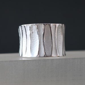 Può includere: Un anello a fascia larga in argento con un designo testurizzato e ondulato. L'anello è realizzato in argento sterling e ha una finitura lucida.