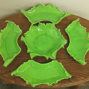 May include: Set of five green ceramic bowls with a unique, ruffled edge design. The bowls are arranged on a wooden surface.