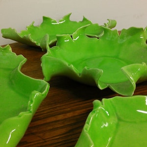 May include: Five green ceramic bowls with a unique, irregular, leaf-like shape. The bowls are arranged on a wooden surface.
