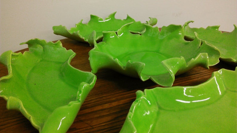 May include: Five green ceramic bowls with a unique, irregular, leaf-like shape. The bowls are arranged on a wooden surface.
