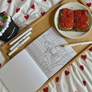 May include: A wooden tray holds a coloring book, markers, and a plate of avocado toast. The coloring book features a line drawing of a person eating pasta. Markers are in a black case and scattered on the tray.