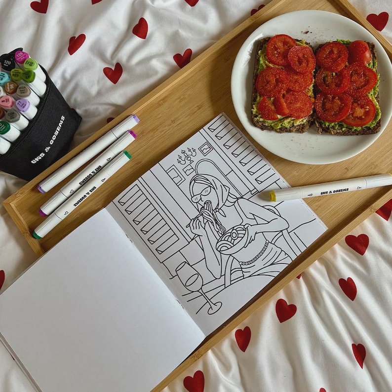 May include: A wooden tray holds a coloring book, markers, and a plate of avocado toast. The coloring book features a line drawing of a person eating pasta. Markers are in a black case and scattered on the tray.