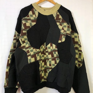 Może przedstawiać: Sweter w stylu vintage z patchworkiem o unikalnym wzorze. Sweter ma czarną bazę z geometrycznymi wzorami w odcieniach brązu, zieleni i beżu. Ma prążkowany kołnierz i mankiety i wisi na drewnianym wieszaku.