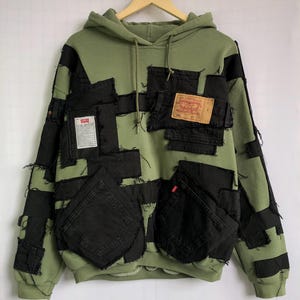 Op de afbeelding: Een olijfgroene hoodie met zwarte denim patches en zakken. De hoodie heeft een capuchon met trekkoord en een Levi Strauss & Co. label. Het ontwerp omvat rauwe randen en een unieke patchwork esthetiek.