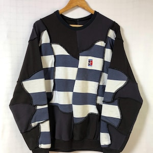 Op de afbeelding: Een patchwork sweatshirt in zwart, marineblauw en wit, hangend aan een houten hanger. Het ontwerp heeft een dambordpatroon en een klein logo. De sweatshirt heeft een ronde hals en lange mouwen.