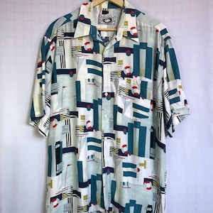 Op de afbeelding: Een overhemd met korte mouwen en knoopsluiting, voorzien van een geometrische nautische print in petrol, marineblauw, wit en rood. Het patroon bevat reddingsboeien, zeilboten en verticale strepen. Het heeft een puntige kraag en een borstzak.