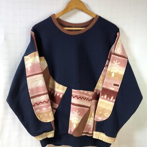 May include: Navy blue sweatshirt with brown trim and patterned patches in pink, beige, and brown. The patches feature geometric and floral designs. The sweatshirt is hanging on a wooden hanger.