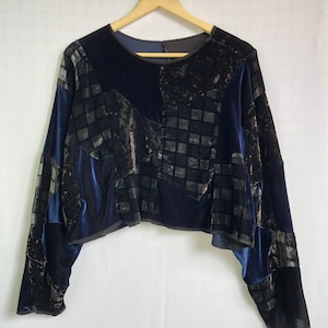 Puede incluir: Top de terciopelo azul oscuro con mangas largas. El top presenta un diseño de patchwork con patrones cuadrados y geométricos en tonos azules y negros. La prenda está colgada de una percha de madera.