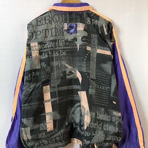 Op de afbeelding: Vintage Nike trainingsjack met een zwart, grijs en perzik printontwerp. De jas heeft paarse en oranje accenten, een Nike-logo en de tekst "AERO OPTIN". De jas hangt aan een houten kleerhanger.