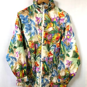 Może przedstawiać: Kurtka w stylu vintage z kwiatowym nadrukiem, wysokim kołnierzem i białym zamkiem błyskawicznym. Kurtka ma żywy wzór niebieskich, fioletowych, czerwonych i żółtych kwiatów z zielonymi liśćmi. Kurtka wisi na drewnianym wieszaku.
