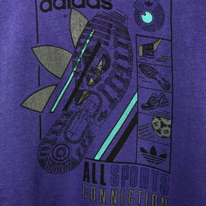 Puede incluir: Camiseta morada con un dise&ntilde;o gr&aacute;fico. El dise&ntilde;o incluye el logotipo de Adidas y la palabra "adidas". Tambi&eacute;n es visible el texto "ALL SPORTS CONNECTION", junto con im&aacute;genes de equipamiento deportivo.