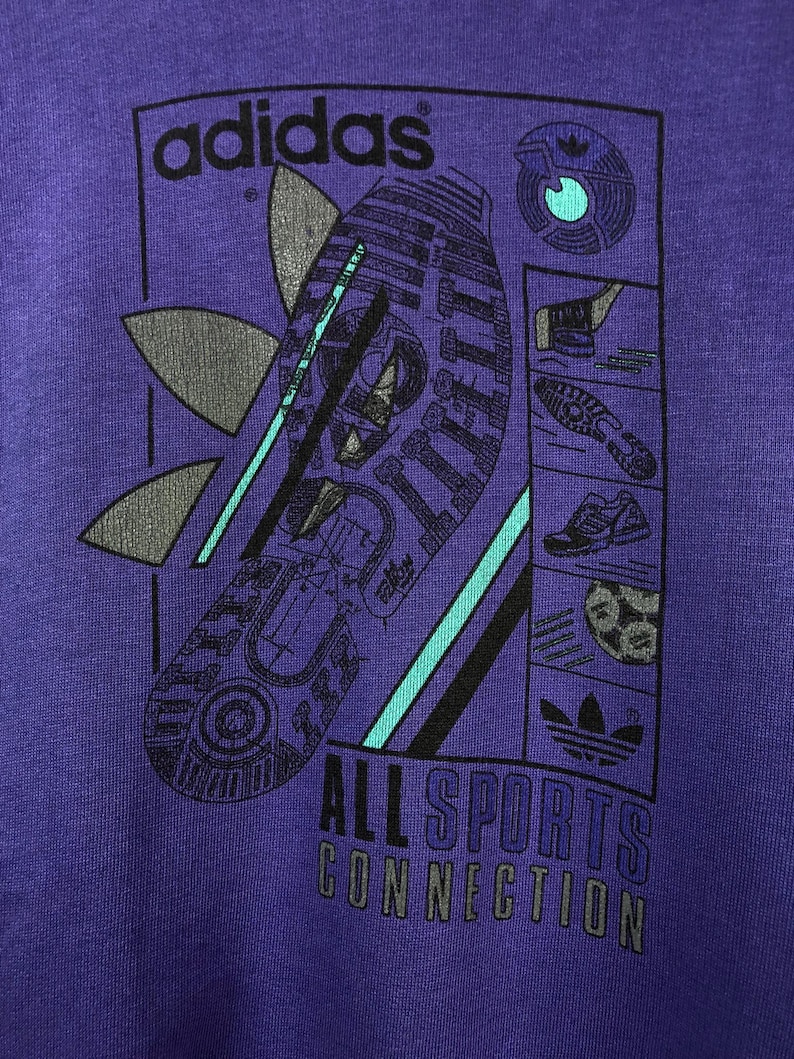 Puede incluir: Camiseta morada con un dise&ntilde;o gr&aacute;fico. El dise&ntilde;o incluye el logotipo de Adidas y la palabra "adidas". Tambi&eacute;n es visible el texto "ALL SPORTS CONNECTION", junto con im&aacute;genes de equipamiento deportivo.