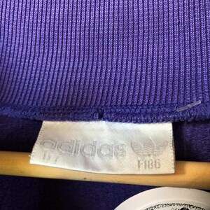 Puede incluir: Primer plano de una tela morada acanalada con una etiqueta blanca. La etiqueta presenta el logotipo de Adidas y la palabra "adidas", adem&aacute;s de los n&uacute;meros "D7" y "F186".