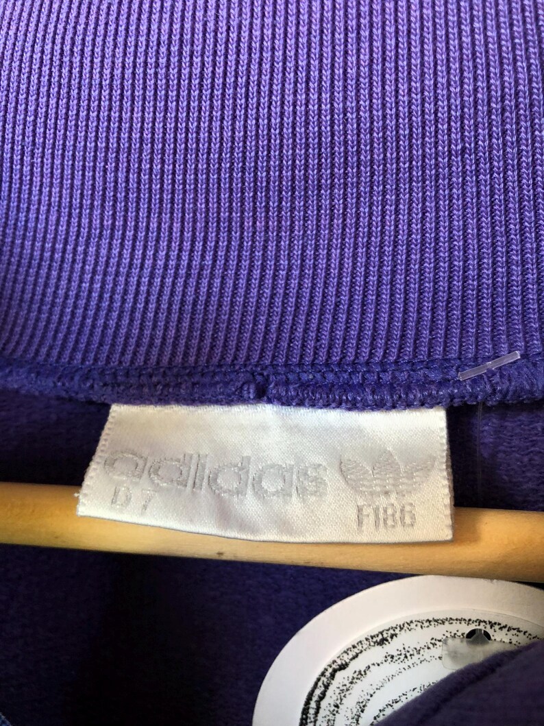 Puede incluir: Primer plano de una tela morada acanalada con una etiqueta blanca. La etiqueta presenta el logotipo de Adidas y la palabra "adidas", adem&aacute;s de los n&uacute;meros "D7" y "F186".
