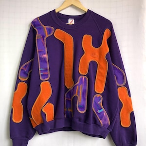 Può includere: Una felpa viola con forme astratte arancioni e viola. La felpa ha un girocollo e maniche lunghe. Le forme arancioni sono delineate con cuciture arancioni. Il logo Adidas è visibile su una delle forme arancioni.