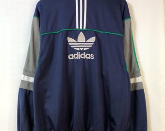 May include: Navy blue Adidas track jacket with white stripes and the Adidas logo. The jacket has gray sleeves with white and green accents. The Adidas logo and text are in white. The jacket is hanging on a wooden hanger.