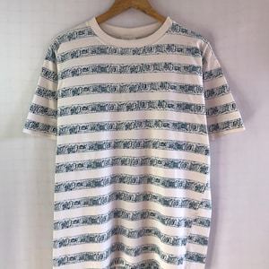 Peut inclure: T-shirt blanc cassé à manches courtes, orné de rayures horizontales bleu-vert. Les rayures présentent un motif géométrique répétitif. Le t-shirt est suspendu à un cintre en bois sur fond blanc uni.