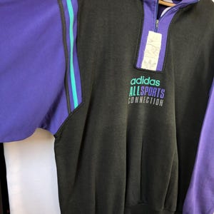 Puede incluir: Sudadera vintage Adidas Allsports Connection en gris carb&oacute;n, morado y verde azulado. La sudadera presenta un cuello con cremallera de cuarto, mangas moradas y rayas verde azulado. El logotipo y el texto de Adidas est&aacute;n impresos en la parte delantera.