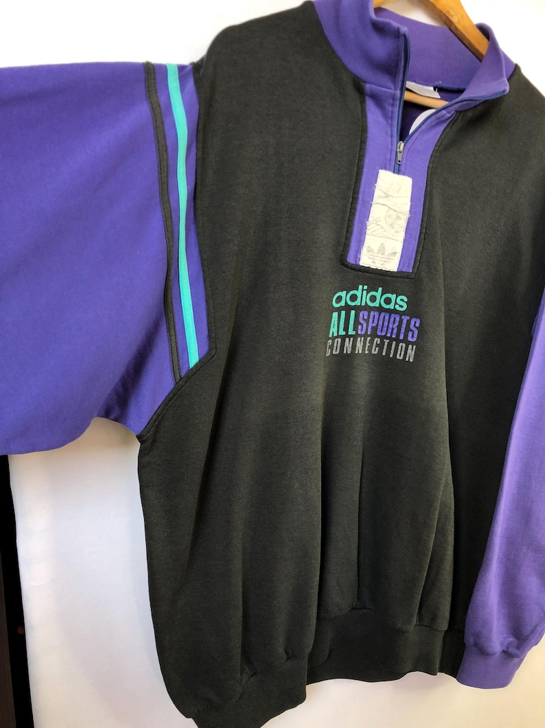 Puede incluir: Sudadera vintage Adidas Allsports Connection en gris carb&oacute;n, morado y verde azulado. La sudadera presenta un cuello con cremallera de cuarto, mangas moradas y rayas verde azulado. El logotipo y el texto de Adidas est&aacute;n impresos en la parte delantera.