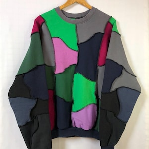 Può includere: Una felpa patchwork in una varietà di colori, tra cui verde, rosa, grigio e blu navy. La felpa ha un girocollo e maniche lunghe. Le toppe sono cucite insieme con cuciture nere. L'indumento è appeso a una gruccia di legno.