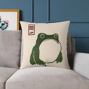 May include: A cream-colored throw pillow with a green frog illustration and a red Japanese character "開" printed on the top left corner.