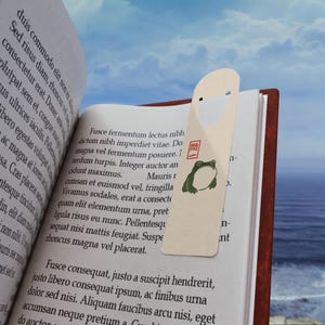 Puede incluir: Un marcapáginas beige con una ilustración de una rana verde y texto japonés está insertado en un libro. El libro está abierto, mostrando texto impreso. El fondo muestra un cielo azul y agua. El diseño es sencillo.