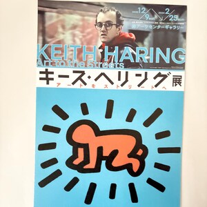 May include: A poster advertising a Keith Haring exhibition titled "Art to the Streets." The poster features a portrait of Keith Haring and a red stick figure with a halo of black lines on a blue background. The text "Keith Haring Art to the Streets" is in white letters on a black background. The text "キース・ヘリング展" is in Japanese characters on a black background.