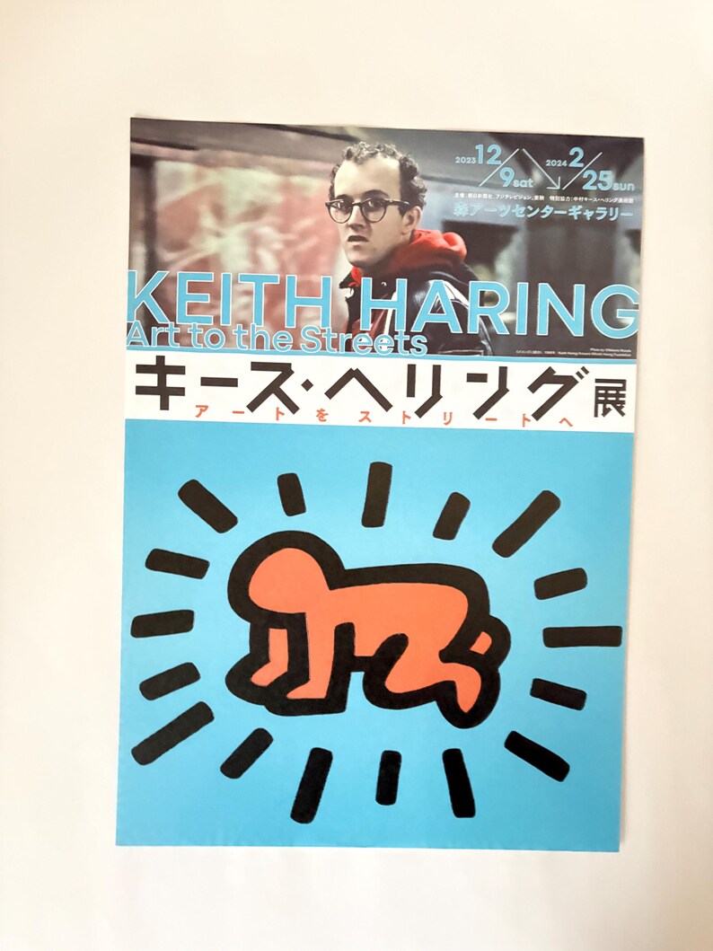 May include: A poster advertising a Keith Haring exhibition titled "Art to the Streets." The poster features a portrait of Keith Haring and a red stick figure with a halo of black lines on a blue background. The text "Keith Haring Art to the Streets" is in white letters on a black background. The text "キース・ヘリング展" is in Japanese characters on a black background.