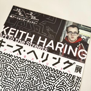 May include: A black and white poster advertising a Keith Haring exhibition titled "Art to the Streets: Keith Haring Exhibition". The poster features a black and white illustration of Haring's signature style, with a close-up of Haring's face in the top right corner.