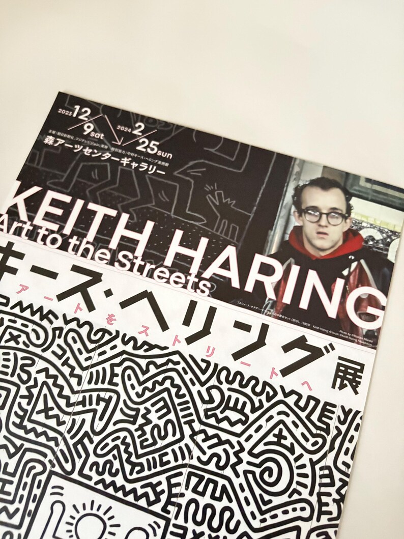 May include: A black and white poster advertising a Keith Haring exhibition titled "Art to the Streets: Keith Haring Exhibition". The poster features a black and white illustration of Haring's signature style, with a close-up of Haring's face in the top right corner.