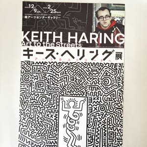 May include: A black and white poster advertising an exhibit of Keith Haring's art. The poster features a black and white illustration of a person with a halo and a series of circles around their torso. The text on the poster reads "Keith Haring Art to the Streets" and "キース・ヘリング展".