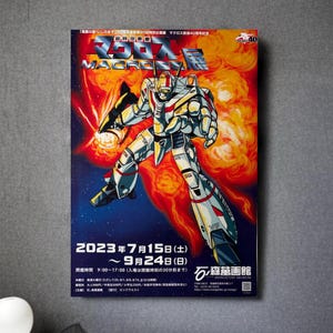 Puede incluir: Un póster que anuncia una exposición de "Macross" en el Museo Mangattan. El póster presenta un robot rojo, blanco y azul sobre un fondo de fuego. La exposición se lleva a cabo del 15 de julio al 24 de septiembre de 2023.