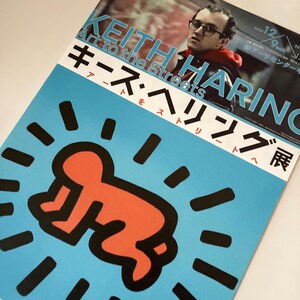 May include: A blue poster with a black and red illustration of a baby in a circle of black rays. The poster advertises a Keith Haring exhibition titled "Art to the Streets" with the text "Keith Haring Art to the Streets" in white and "キース・ヘリング展" in red.
